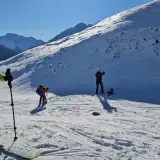 Un grupo de esquiadores se prepara para el esqui extremo en una ladera nevada, con picos montañosos al fondo.