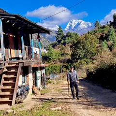 Hombre con bastón de trekking en un camino rural, con una casa tradicional y montañas nevadas al fondo. Una aventura de trekking Himalaya.
