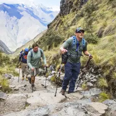 Tres excursionistas suben un sendero rocoso en las montañas, usando bastones de trekking para ayudarse.