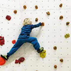 Niño rubio con camiseta a rayas azules y pantalón azul practica escalada para niños en un rocódromo blanco con presas de colores.