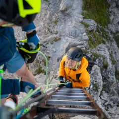 Dos personas con equipo ferrata escalan una pared rocosa, una subiendo una escalera metálica y la otra asegurándola.