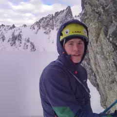 Alpinista con casco amarillo y ropa de montaña, rodeado de picos nevados y niebla.