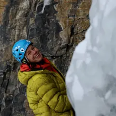 Inés Papert, sonriendo, con casco azul y chaqueta amarilla, lista para escalar en un acantilado rocoso y helado.