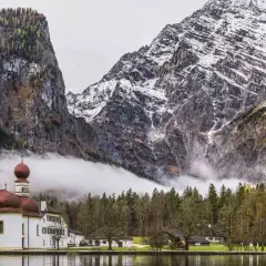 Iglesia blanca con cúpulas rojas junto a un lago, rodeada de bosques y majestuosas montañas nevadas en los Alpes alemanes.
