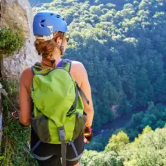 Mujer con casco y mochila verde en vía ferrata, con vistas a un bosque frondoso. Afortunadamente, no hubo ningún accidente de escalada.