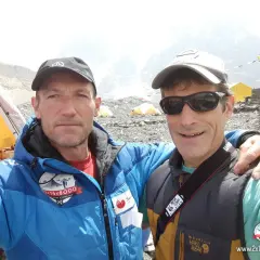 Alberto Zerain y un compañero posan sonrientes en un campamento de montaña, con tiendas amarillas y banderas de oración al fondo.