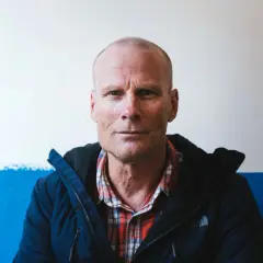 Retrato de Alex Lowe, un hombre calvo con camisa de cuadros y chaqueta azul, mirando fijamente a la cámara.