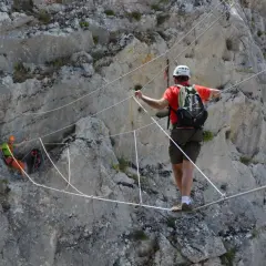 Un hombre con casco y mochila cruza un puente de cables en una pared rocosa. Otro excursionista, con casco naranja, se ve en la distancia. Esto es una demostración de que es rapel.