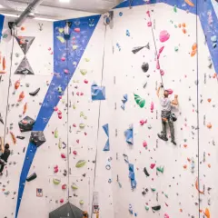 Escaladores en rocódromo, uno asciende con un asegurador automático de escalada, el otro en la pared azul.