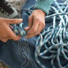 Manos preparan cuerda para machard escalada. Botas de montaña y lona azul con nudos dibujados.