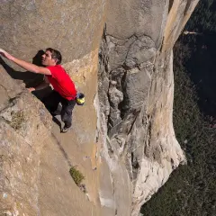 Un escalador en solitario asciende una pared de roca imponente, capturando la esencia de un documental de escalada. La vista es vertiginosa, con un bosque denso abajo.