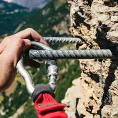 Mano enguantada se engancha a la **línea de vida vía ferrata** en una pared rocosa, con un paisaje montañoso al fondo.