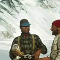 Dos montañistas sonríen en la nieve. Uno, con gorra y camisa de cuadros, sostiene un pico. El otro, con gorro rojo, parece ser Tom Hornbein.