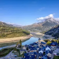 Pueblo de piedra junto a un embalse, con montañas nevadas de los picos de Huesca al fondo.