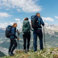 Tres excursionistas con mochilas y bastones admiran un paisaje montañoso. Sus prendas, ideales para distintos tipos de goretex, los protegen del clima.