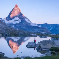 Persona sentada en roca junto a lago, contemplando el reflejo del **Cervino** al amanecer.