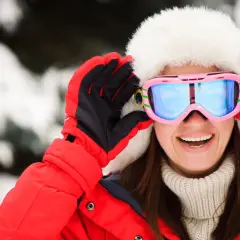 Mujer sonriente con gorro de piel y gafas de esquí rosas, lista para la nieve. Explora las categorías de gafas de esquí para encontrar tu estilo.