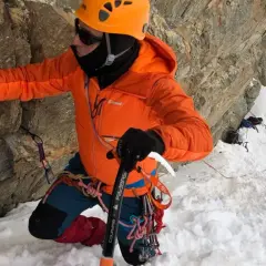 Alpinista con casco naranja y chaqueta brillante usa un piolet para escalar una pared rocosa nevada.