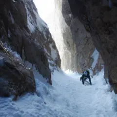 Alpinista asciende por un estrecho cañón nevado, usando piolet y crampones. La estaca de nieve se ve firme bajo sus pies.