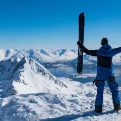 Persona en la cima de una montaña nevada, sosteniendo un splitboard que es un equipo versátil para ascender y descender.