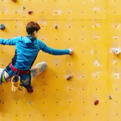 Una escaladora se enfrenta a una pared amarilla, buscando el siguiente agarre en una escalada de dificultad.