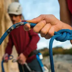 Manos atando nudos de escalada con una cuerda azul. En el fondo, otro escalador con casco.