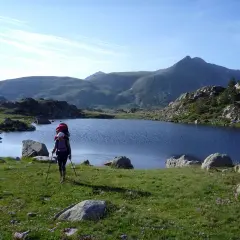 Senderista con mochila y bastones camina por un prado verde hacia un lago azul en los Pirineos de GRP Andorra.