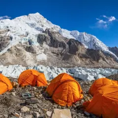 Campamento de expedición en la base de la cordillera más alta del mundo, con tiendas naranjas y glaciares imponentes bajo un cielo azul.