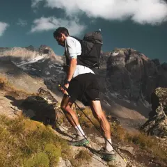 Kilian Jornet asciende una ladera rocosa en los Pirineos, con bastones y mochila, bajo un cielo azul con nubes.
