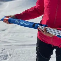Persona con chaqueta rosa sostiene una arva, sonda y pala ORTOVOX en una bolsa azul, lista para la aventura en la nieve.