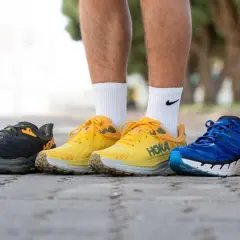 Cinco pares de zapatillas Hoka con gran amortiguación, en colores azul, gris, amarillo, azul y naranja, alineados en un camino.