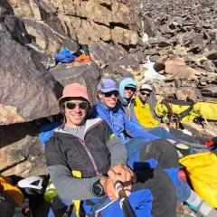 Cuatro montañistas sonríen en la ladera rocosa, listos para la aventura. El **bru busom** de uno de ellos, un sombrero rosa, destaca entre el equipo de expedición.