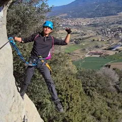 Hombre con casco azul y arnés, asegurado a un cable de **material vía ferrata**, disfruta de las vistas del valle.