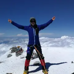 Javier García Díaz, alpinista, celebra en la cima nevada, rodeado de nubes y un cielo azul intenso.