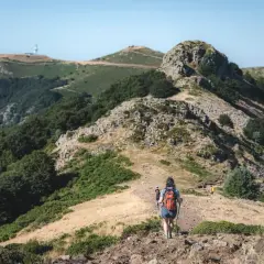 Senderismo en montaña con bastones, comparando rutas en AllTrails vs. Wikiloc. Paisaje verde y rocoso bajo un cielo azul.