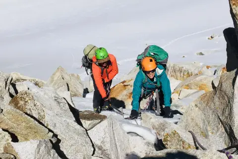 Dos escaladores con mochilas y cascos navegan por rocas y nieve en la montaña. Sus aventuras son como libros abiertos de la naturaleza.