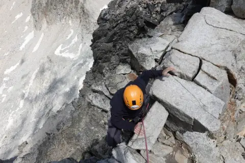 Alpinista con casco naranja escala roca, buscando el aneto por coronas.