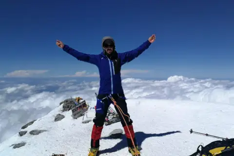 Javier García Díaz, alpinista, celebra en la cima nevada, rodeado de nubes y un cielo azul intenso.