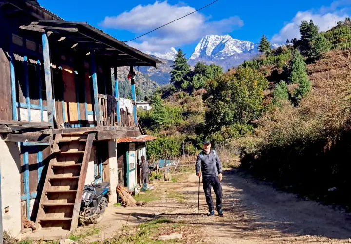Hombre con bastón de trekking en un camino rural, con una casa tradicional y montañas nevadas al fondo. Una aventura de trekking Himalaya.