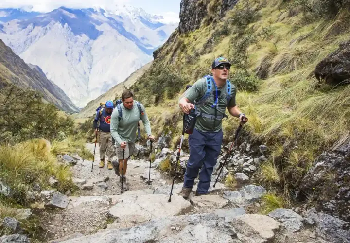 Tres excursionistas suben un sendero rocoso en las montañas, usando bastones de trekking para ayudarse.
