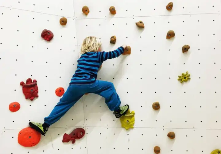 Niño rubio con camiseta a rayas azules y pantalón azul practica escalada para niños en un rocódromo blanco con presas de colores.