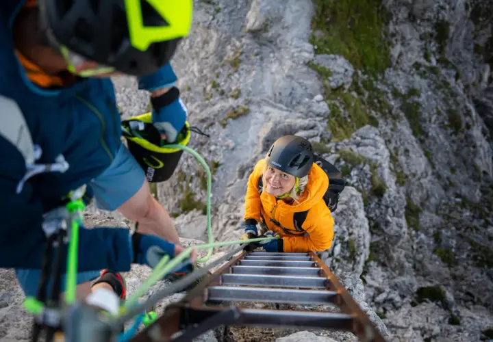 Dos personas con equipo ferrata escalan una pared rocosa, una subiendo una escalera metálica y la otra asegurándola.