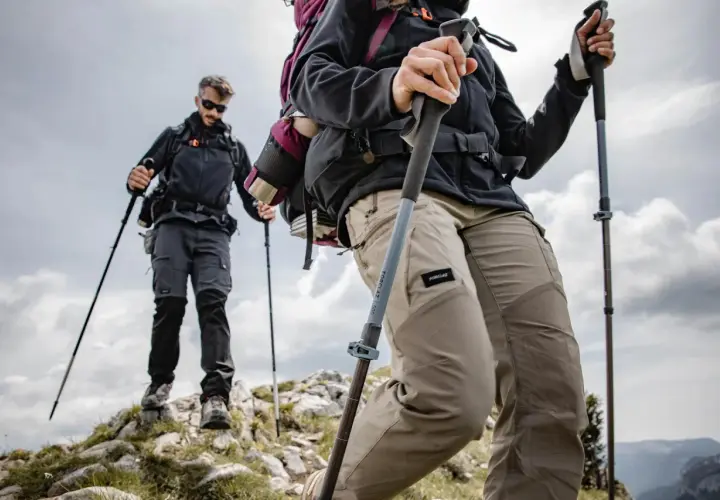 Dos excursionistas suben una ladera rocosa, usando sus pals per caminar para mayor estabilidad. El cielo nublado sugiere una aventura desafiante.