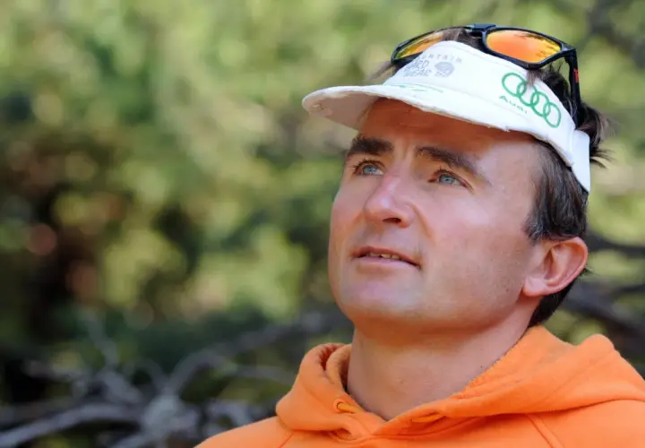 Ueli Steck, con gorra blanca y sudadera naranja, mira hacia arriba con determinación.