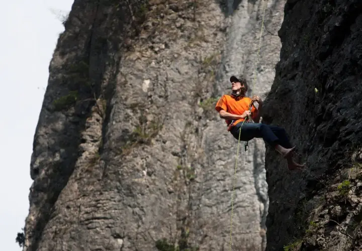 Dean Potter desciende por una pared rocosa, con el sol brillando sobre su camiseta naranja.