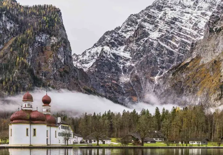 Iglesia blanca con cúpulas rojas junto a un lago, rodeada de bosques y majestuosas montañas nevadas en los Alpes alemanes.