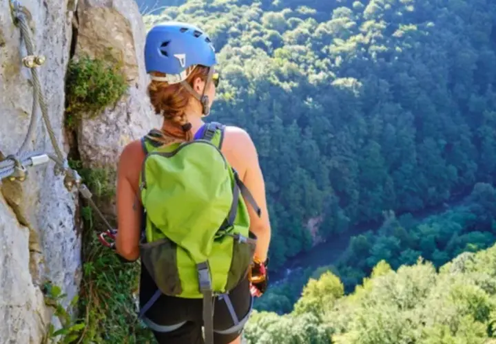 Mujer con casco y mochila verde en vía ferrata, con vistas a un bosque frondoso. Afortunadamente, no hubo ningún accidente de escalada.