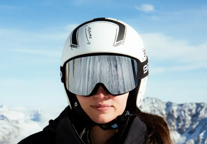 Mujer con casco de esquí blanco y gafas espejadas, lista para deslizarse por la montaña.