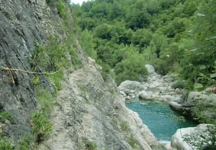Sendero rocoso con cuerda de seguridad a lo largo de la pared de la garganta de Escuain, con un río turquesa y vegetación exuberante al fondo.