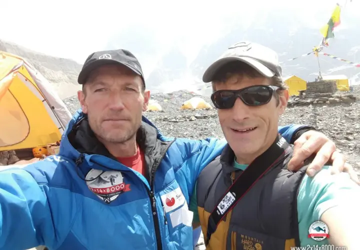 Alberto Zerain y un compañero posan sonrientes en un campamento de montaña, con tiendas amarillas y banderas de oración al fondo.